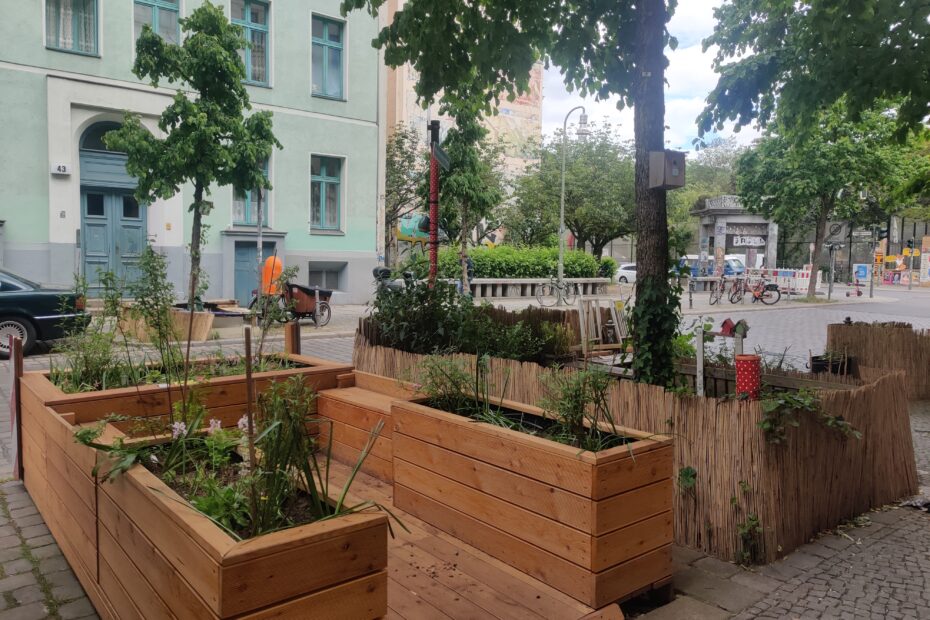 Vom Bürgersteig aus sieht man in Richtung Straße zwei große Parklets, mit Sitzbänken aus Holz, bepflanzten Blumenkübeln und Baumscheiben. Die Sraße im Hintergrund ist leer. So könnte ein zukünftiger Kiezblock in Friedrichshain-Kreuzberg aussehen.