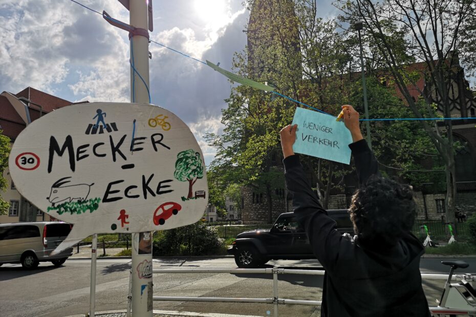 Draußen an einer Straße hängt an einer Laterne ein Schild mit der Aufschrift "Meckerecke". Auf dem Schild sind kleine Symbole, unter anderem ein Baum, ein Auto und ein Zebrastreifen. Rechts daneben hängt eine Person einen Zettel mit der Aufschrift "weniger Verkehr" an eine improvisierte Wäscheleine.