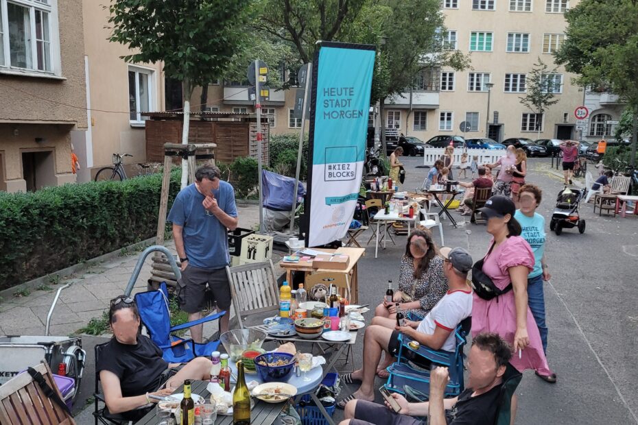 Foto vom Nachbarschaftsfest: Im Vordergrund ein langer gedeckter Tisch um den Menschen sitzen, im Hintergrund auf der Straße spielende Kinder, Häuser und Bäume, keine Autos